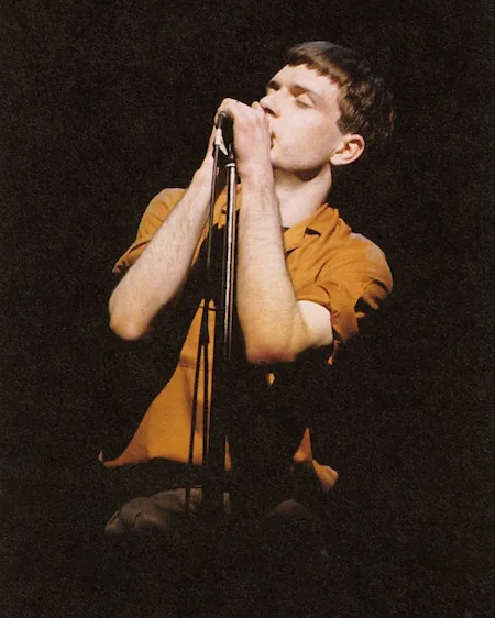 Ian Curtis cantando frente al micrófono con una camisa mostaza durante un concierto