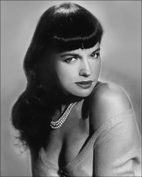 Retrato en blanco y negro de la pin-up Bettie Page, con su chasquilla corta, un collar de perlas y un chaleco caído sobre el hombro