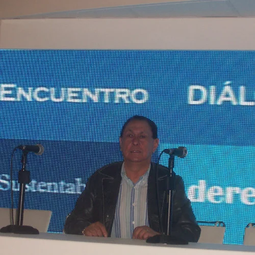 Carlos Quiroga hablando de pie ante un atril, con chaqueta de cuero, frente a una pantalla en un encuentro