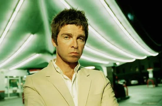 Noel Gallagher con chaqueta clara frente a una gasolinera iluminada de noche
