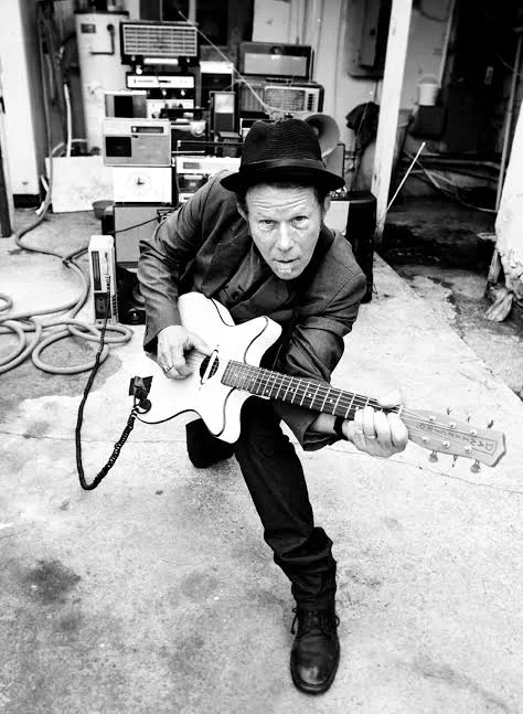 Tom Waits agachado con una guitarra, de sombrero, frente a una pila de amplificadores antiguos